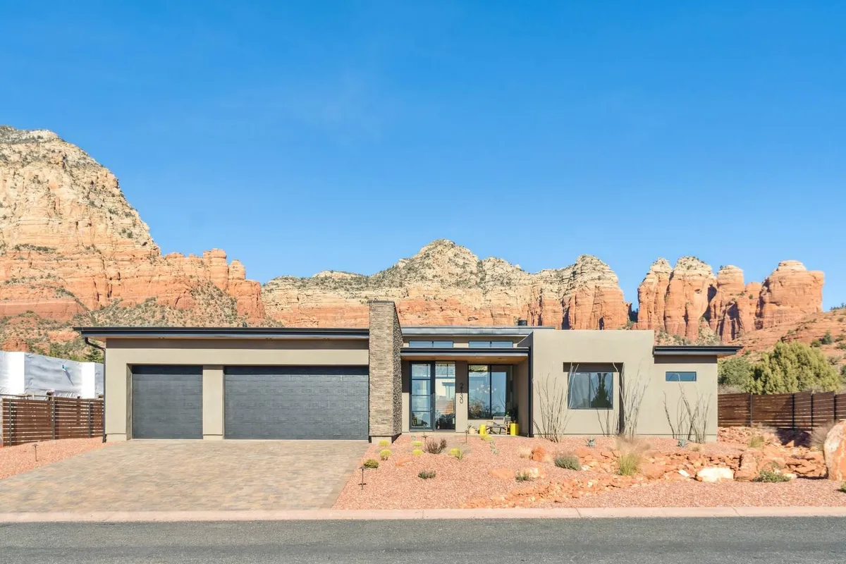 The image showcases a sleek, modern single-family home set against the dramatic backdrop of Sedona’s iconic red rock formations. Expansive glass windows reflect the vibrant sunset hues, while a beautifully landscaped yard invites relaxation and gatherings. The serenity of the scene is enhanced by the open skies, ideal for evening stargazing.