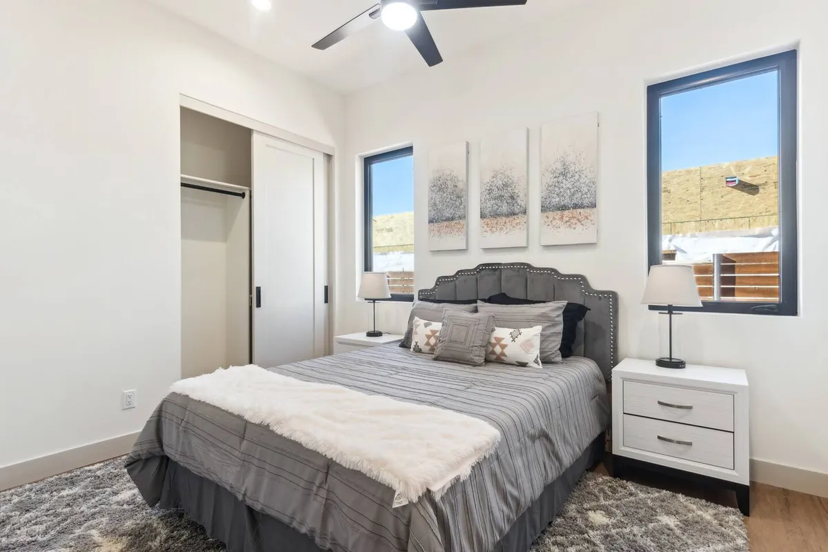 A cozy, modern bedroom featuring a bed with decorative pillows, artwork above the headboard, and nightstands topped with lamps. Sunlight pours in through windows, illuminating the soft linens and neutral décor. The overall feeling is restful and inviting, with a sense of tranquility ideal for unwinding after a day of adventure.
