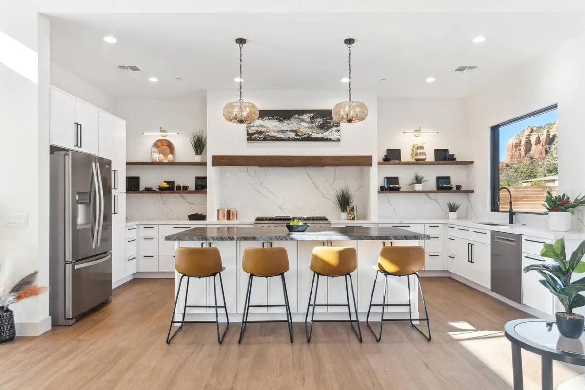 A spacious kitchen featuring a large island at the center, illuminated by modern pendant lights. The minimalist cabinetry and natural stone backsplash complement the inviting atmosphere, while a window with a view brings in natural light. This scene frames the kitchen as a social and functional space for both cooking and gathering.
