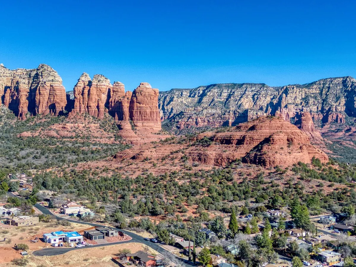 A sweeping view of Sedona’s iconic red rock formations framed by lush green vegetation below, with a small residential area tucked into the landscape. The natural beauty is bathed in golden sunrise or sunset light, emphasizing the majestic geological features.