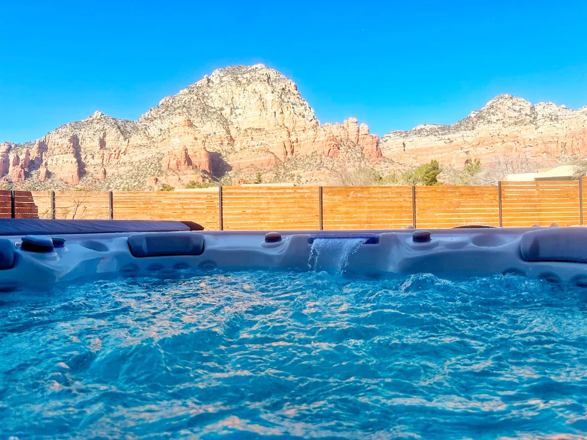 This exterior image centers on a hot tub positioned to take advantage of sweeping mountain views beneath a clear blue sky. The hot tub is nestled in a private outdoor area, surrounded by natural landscape and distant peaks, evoking tranquility and relaxation. The scene suggests people relaxing in the tub while enjoying the expansive scenery, ideal for sunset soaks or stargazing.