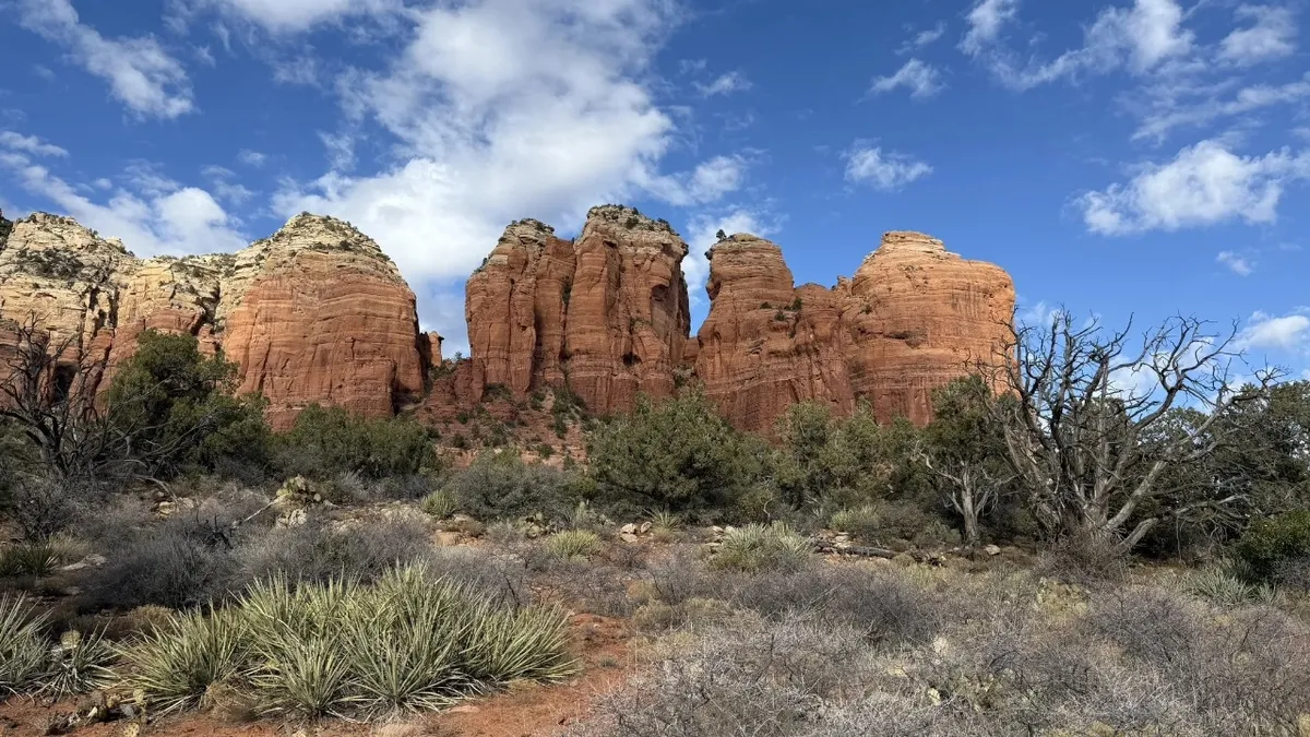 Dramatic red rock formations rise against a backdrop of distant mountains and desert vegetation. The scene is awe-inspiring, highlighting both the rugged terrain and the unique colors of Sedona’s landscape, perfect for hiking and photography.